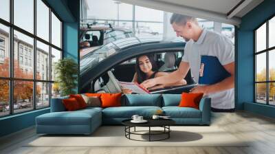 Nice consultant stands at black car and points on paper. He holds plastic tablet in other hand. Nice and young woman sits in car and looks at document. She is concentrated. Wall mural