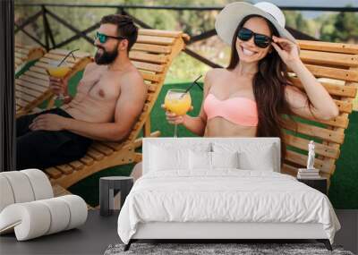 Nice picture of girl and guy sitting on sunbeds. She looks on camera and smiles. Girl touches her sunglasses. She poses. Guy looks forward. He holds glass of cocktail. Wall mural