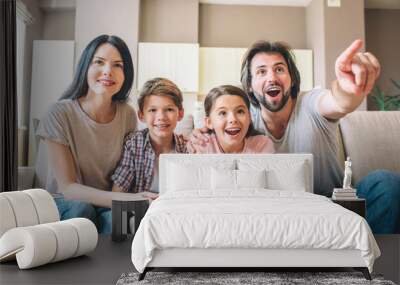 Parents are sitting together with their kids on sofa. Children play the game on playstation. Guy is pointing forward and screaming. Woman helps to play her son. Wall mural