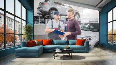 Picture of two young men stand in garage together. Bearded guy smiles and points on plastic tablet with documents. Man in grey uniform and cap talk and look at customer. He is serious. Wall mural