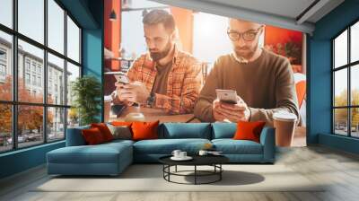 Take a look on beautiful and handsome bearded men sitting at te table and holding phones. They are looking only at their screens. Guys are busy. Close up. Wall mural