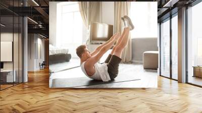 Young well-built man go in for sports in apartment. He lying on back and reach feet with hands. Young man stretching. Yoga exercise. Wall mural