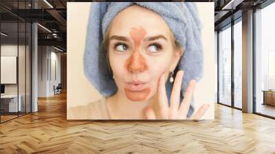 A beautiful girl with a towel on her head makes a multi mask on the t-zone of orange color with a scrub. The cute blonde is having fun doing a home care, moisturizing and smoothing facial mask Wall mural