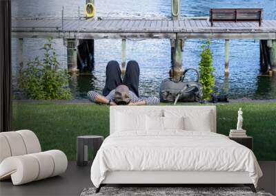 A person in casual attire rests face down on the grass beside a calm lake, with a jetty and bench in the background. Wall mural