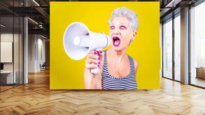 Senior grey haired woman screaming loudly in a megaphone on yellow background demonstrating Wall mural