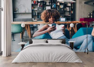 young black woman drinking and chatting with smartphone in modern loft Wall mural