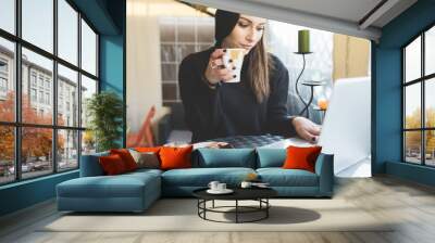 young handsome caucasian woman sitting in a bar working with her laptop, drinking from a cup, lookin Wall mural