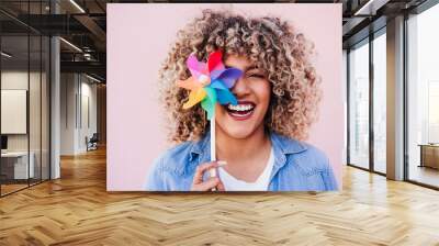 beautiful happy hispanic woman with afro hair holding colorful pinwheel. pink background,wind energy Wall mural