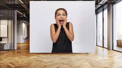 Young beautiful little girl covers her mouth in shock, emotionally looks with great surprise at the camera, opens widely eyes and mouth on a white isolated background. Wall mural