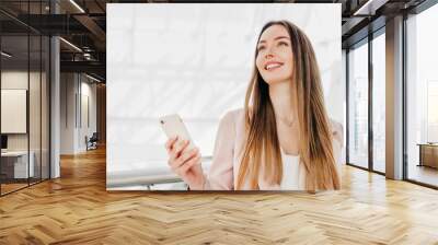 Business woman stands indoors in an office building holding a mobile phone and smiling Wall mural