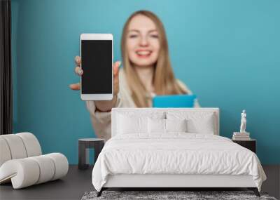 Happy student girl shows a blank screen of a mobile phone at the camera and smiles, girl in blur. Isolated on a blue background in the studio. Mockup Wall mural