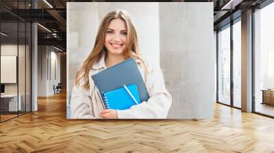 Young blonde girl student smiling against university. Cute girl student holds folders and notebooks in hands, copy space. Learning, education concept Wall mural