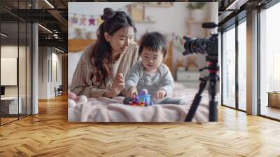 a mother and her child sitting on the bed, playing with toys while being filmed for an online video content production. An Asian mother and her young child play together on a bed Wall mural