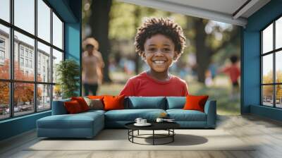 A young boy wearing a red shirt is smiling and standing in a park. There are other children in the background, some of them playing and others watching. Concept of happiness and playfulness Wall mural