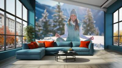 an attractive woman in her late thirties, doing yoga on the snow covered mountain with a bright blue sky and pine trees in the background. Wall mural