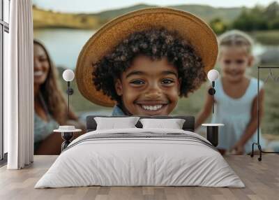 Happy young Black boy wearing a straw hat at a lakeside picnic, friends around. Wall mural