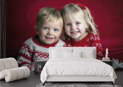 Two Caucasian toddlers, a boy and a girl, smiling in festive sweaters, sitting against a red backdrop. Wall mural