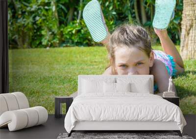 Little girl sunbathes in the sun lying on green grass near palm tree in big slippers. Wall mural