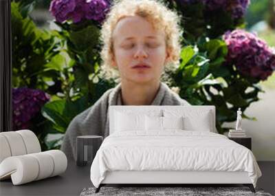 Portrait of a girl of European appearance with blond curly hair, meditates in nature, hands clasped in namaste, against the background of hydrangea flowers. Wall mural