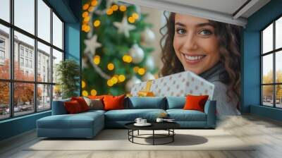 Portrait of attractive young woman holding Christmas present in her hands against background of New Year tree with decorations and burning garlands. Wall mural