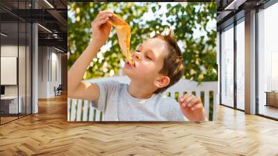 boy eating pizza. child closed his eyes in pleasure and takes a bite of pizza piece holding it aloft Wall mural