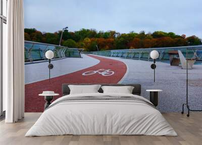 Low-angle view of winding red color bike lane with white bicycle sign on the New Pedestrian Bridge in Kyiv, Ukraine. Beautiful autumn colored trees in the background Wall mural