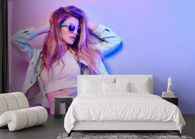 Fashionable hipster woman in Trendy outfit, stylish hair, makeup on pink purple neon light. Blonde model in hoodie, fashion jeans, sunglasses. Beautiful bad sexy girl in neon style Wall mural