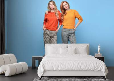 Two Lovable embracing fashionable woman sisters in Trendy orange yellow outfit. Studio shot of Carefree beautiful funny stylish friends smiling on blue. Happy fashion girl, colorful positive mood Wall mural