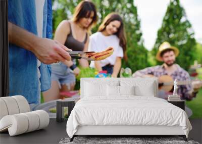 a man's hand holds a barbecue tongs with a juicy delicious meat steak against the background of a barbecue grill with meat and vegetables and a group of friends on a picnic who are having fun and Wall mural