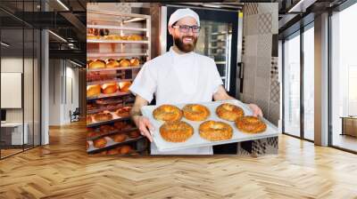 attractive Baker in white uniform holding a tray with freshly baked bagels with sesame and poppy seeds on the background of a bread factory or bakery Wall mural