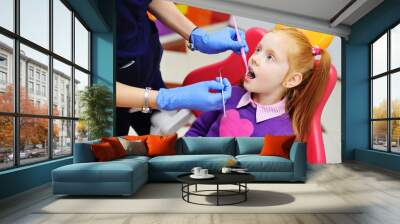 children's dentist examines the teeth and mouth of the child - a cute red-haired girl sitting in a dental chair. Pediatric dentistry Wall mural