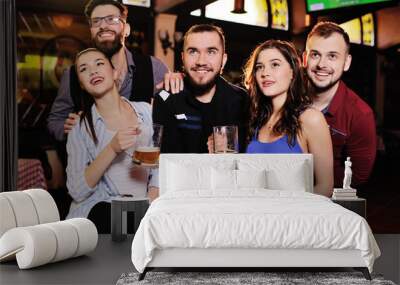 group or company of friends - young guys and girls holding glasses of beer, watching football, laughing and smiling at the bar during the Oktoberfest festival Wall mural