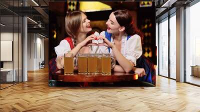 two young attractive girls in Bavarian dresses sitting at a table in a bar or pub drinking beer and clinking glasses during the celebration of the Oktoberfest Wall mural