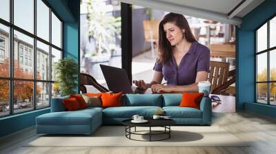 Portrait of beautiful woman working on laptop while sitting in cafe with coffee in paper cup Wall mural