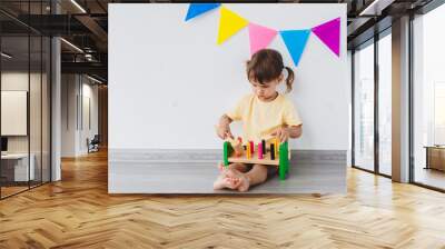 girl 2 years old toddler sits on the floor playing with learning multicolor toy pounding bench at home. on a white background with colored flags, Count learning. activity for preschoolers Wall mural