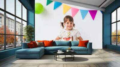 smiling white toddler girl 2 years old sitting in high chair on white background with colorful flags holding piece of birthday cake, looking at camera, holiday concept Wall mural