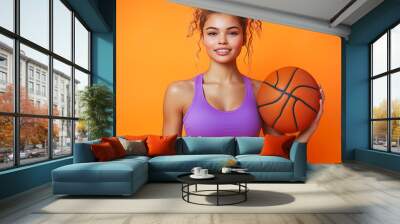 Young, athletic fitness trainer woman in sportswear holding a basketball, isolated on a plain orange background Wall mural