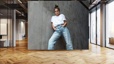 A beautiful young woman in full growth stands against the wall. A stylish girl in blue jeans and a white T-shirt stands against the background of a gray concrete wall. 80's 90's style Wall mural