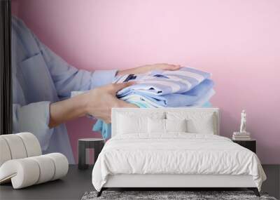 Close up of young woman wearing blue cotton shirt holding stack of perfectly folded multicolor shirts. Female w/ pile of different color clothing in her hands on pale pink background. Laundry concept. Wall mural