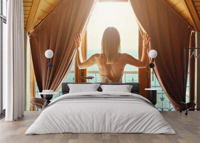 New day new life concept. Young attractive woman wearing nighties opens curtains of big windows with sea view lets the sunshine in room. Back of fit blonde female in sexy silk nightwear. Background. Wall mural