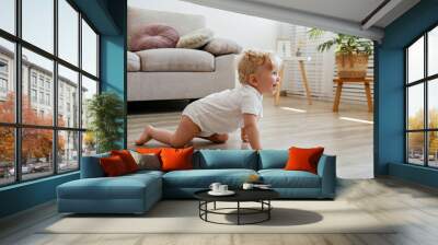 One year old child learning to crawl on the wooden floor of living room. Interior background. Adorable blonde little girl in pink crawlers at home. Close up, copy space for text. Wall mural