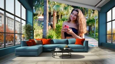 Portrait of young beautiful woman with long wavy brunette hair with her phone in tropical destination park with greenery on the background. Joy of summer holidays concept. Close up, copy space. Wall mural