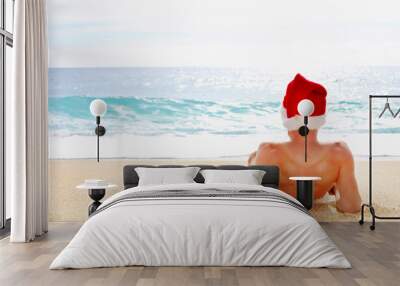 Rear view of fit young man in a Santa hat celebrating Christmas on tropical island sandy beach, enjoying sea view, blue ocean waves on sunny day. Christmas in july concept. Background, copy space. Wall mural