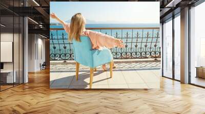 Young attractive woman wearing nighties welcoming new day on balcony of luxurious hotel with sea view. Fit blonde female in sexy silk nightwear sitting in armchair on sunshine. Background, copy space. Wall mural