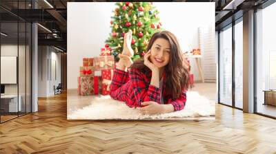 Young beautiful brunette woman in plaid checkered pajamas in decorated bedroom interior with gift box and Chrictmas tree on background. People on Christmas morning concept. Close up, copy space. Wall mural