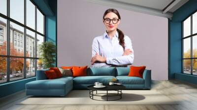 Young beautiful confident businesswoman wearing blue blouse & black frame glasses, standing w/ folded hands & smiling. Attractive brunette female with crossed arms. Background, close up, copy space. Wall mural