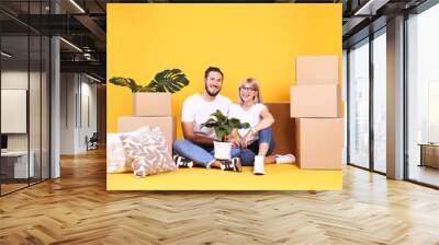 Young married couple moving in new apartment, sitting on the floor between many boxes. New home concept. Bearded man and blonde woman resting over yellow wall. Background, copy space, close up. Wall mural