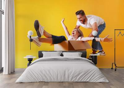 Young married couple moving into a new home. Attractive blonde woman sitting in cardboard box while bearded man pushes her. Newely weds fooling around. Isolated yellow background, copy space, close up Wall mural