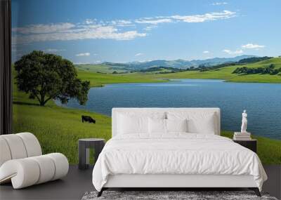 Two cows graze peacefully on vibrant green grass beside a calm lake. Lush hills and distant mountains create a tranquil backdrop under a blue sky Wall mural