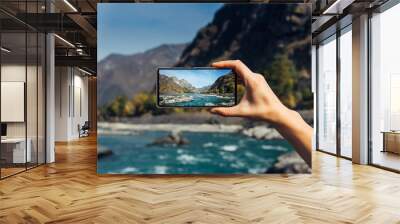 Female hand with smartphone close-up. Taking photo on smartphone while traveling. Delightful mountain landscape, stormy river, rocks and blue sky. Wall mural